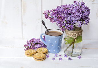 cup of coffee with biscuits and bouquet of lilac on white wooden background