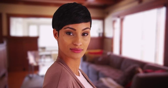 Close up of lovely black female with pixie haircut smiling at camera inside living room. Close-up of African American woman in her 20s looking at camera with confident expression 