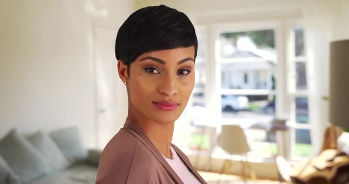 Close up of lovely black female with pixie haircut smiling at camera inside living room, light coming through window. Close-up of African American woman looking at camera with confidently 