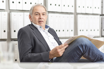 Älterer Mann mit weißem Hemd  sitzt auf einem Stuhl und blättert in einer auf Dokumenten Akte