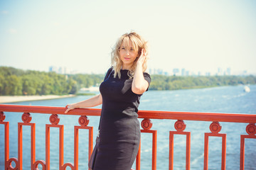 Attractive american blonde hair woman walk in the city wear in small black dress and bag. Lady on a bridge in European town