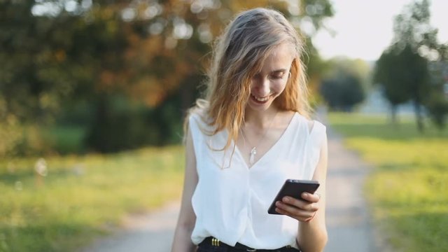 Young happy smiling girl using modern smart phone outdoor attractive woman texting sms message cellphone walking park blurred green trees nature background stylish laughing chatting online technology