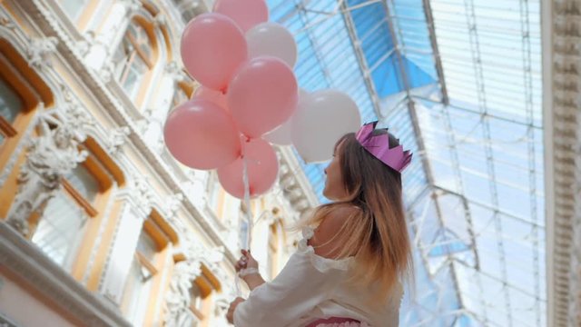 Portrait of Young happy woman with paper crown, colourful white and pink balloons smiling and laughing. Birthday girl in beautiful near old arhitecture. passage. Slow motion.