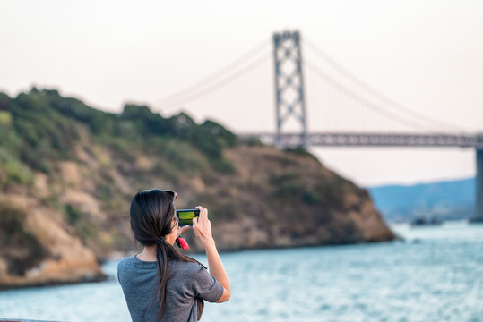 Female tourist photographing San Francisco Bay Bridge at sunset