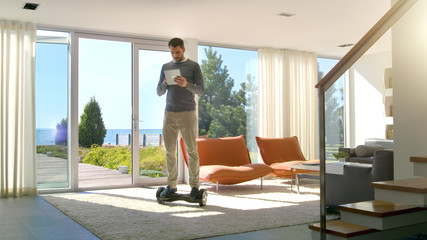 Tech Industry Businessman Working From Home Uses Tablet Computer and Rides Around the House on His Gyro Scooter. His Modern House has Seaside View.