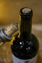A dark bottle of wine with a jammed cork and a bottle opener with a damaged cork on a light brown...