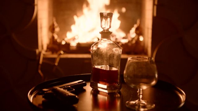 Crystal carafe with whiskey and the glass opposite the fireplace on eve. Pistol on the table