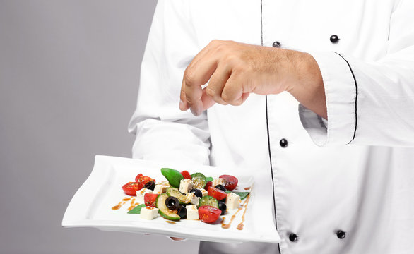 Male Chef In Uniform Adding Seasoning To Salad On Grey Background