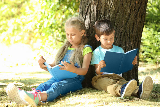 Cute Little Children Reading Books Near Tree In Park