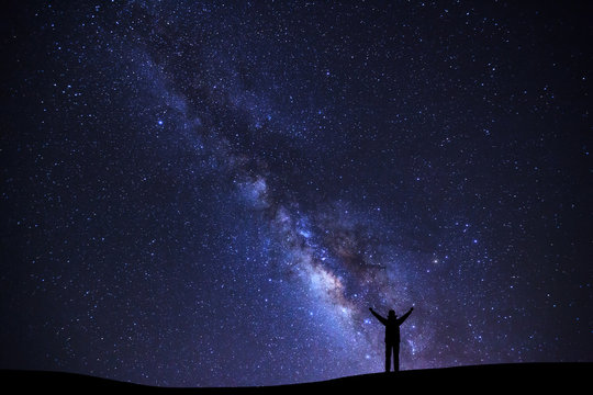 Fototapeta Landscape with milky way galaxy, Starry night sky with stars and silhouette of a standing sporty man with raised up arms on high mountain.