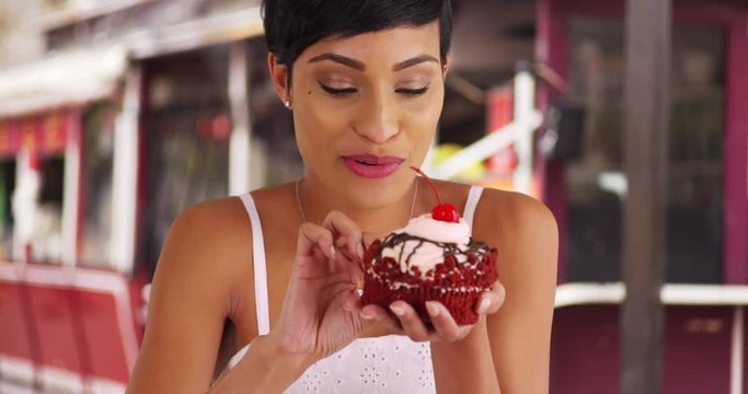 Close up of attractive black female eating big red velvet cupcake outside bakery in summertime. Portrait of woman in her 20s enjoying fancy dessert outdoors, smiling 