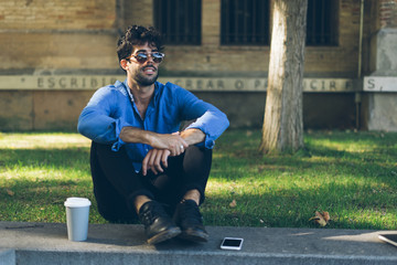 Attractive model look male wearing sunglasses is sitting on a street while having coffee break with a mobile phone lying next to him. Young businessman is smiling a side while enjoying the sunny day.