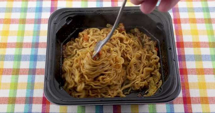 Video of eating beef flavored ramen noodles with a fork in a black microwavable tray atop a colorful place mat.