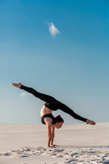 Young woman practicing handstand on beach with white sand and bright blue sky