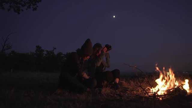 multiethic two girls sitting near bonfire talking enjoying magnificent night under moon with sea on background slow motion
