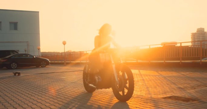 TRACKING Beautiful Caucasian female biker riding her custom built cafe racer motorcycle on a rooftop parking lot, beautiful sunset over city in the background. 4K UHD 60 FPS