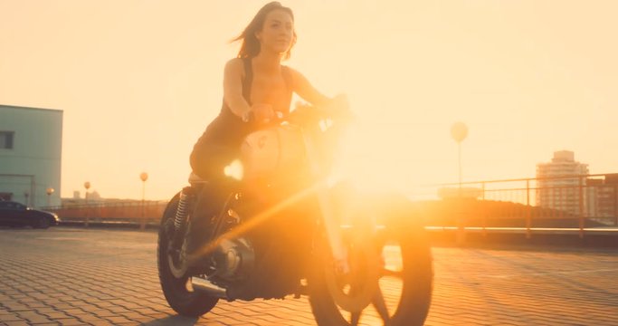 Beautiful Caucasian female biker riding her custom built cafe racer motorcycle on a rooftop parking lot, beautiful sunset over city in the background. 4K UHD 60 FPS