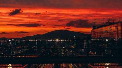 Fototapeta premium Stunning sunset on huge railroad terminal with multiple railway tracks, glass facade on the right and hills ridge in distance; reddish sky, autumn evening, many reflections and lights, Adler, Russia