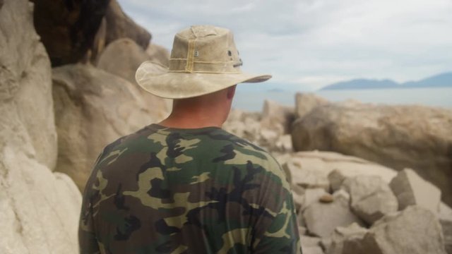 A man explores a rocky terrain in a tracking shot. In front of a man is a beautiful horizon of the ocean and tall mountains.