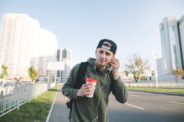 Portrait of a smiling young man with a cup of coffee in his hands, listening to music in headphones against the background of the city and looking at the camera. Lifestyle concept.