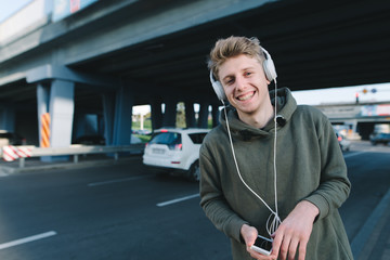 Street portrait of a happy young man who smiles, and listens to a musician in headphones in the background of urban architecture. A look at the camera.