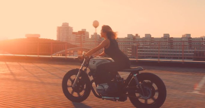 TRACKING Beautiful Caucasian female biker riding her custom built cafe racer motorcycle on a rooftop parking lot, beautiful sunset over city in the background. 4K UHD 60 FPS