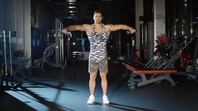 Athletic handsome man works out and lifts hands with dumbbells in gym