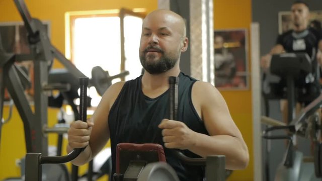 White bald bearded man is doing exercise on hummer row machine in gym