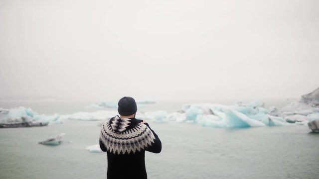 Back view of young traveling man standing in Jokulsalon ice lagoon in Iceland and take photos og glaciers on smartphone.