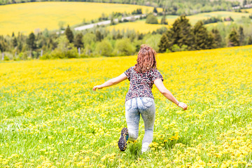 Back of young woman running, jumping in air and smiling on countryside yellow dandelion flower fields in summer grass in Ile D'Orleans, Quebec, Canada back