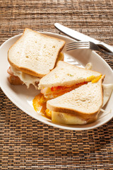 Egg sandwiches with cheese, bacon and tomato. On white plate, with knife and fork on woven placemat.