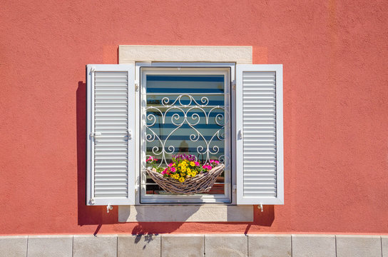 Window with white shutters and beautiful blooming flowers against a red wall