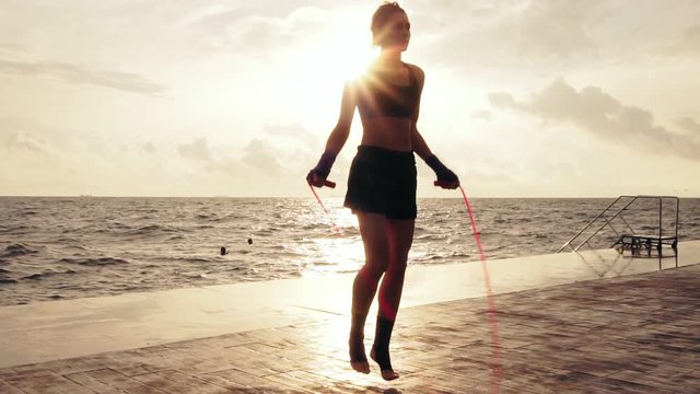 Young woman working out on the jump rope against the sun by the beach in slowmotion. Girl jumping on a skipping rope by the sea