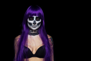 close-up portrait  girl of purple hair (wig)  with terrifying halloween skeleton makeup in black bra and a tattoo on his arm over black background looks at the camera
