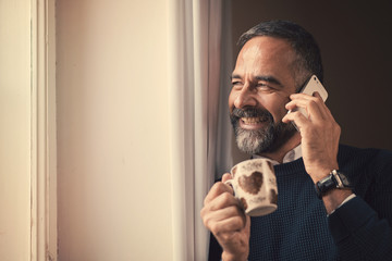 Senior older man talking on his cell phone while having his morning coffee, enjoying a window view