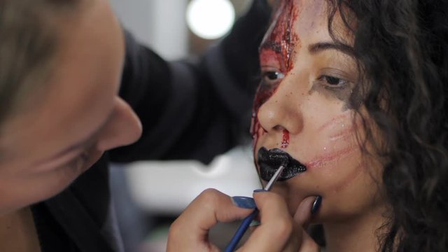 Make-up artist make the girl halloween make upin studio.Halloween face art. Woman applies on black lipstick with brush on lips of latin girl. War-paint with blood, scars and wounds.Slow motion.