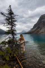 Woman in a dress standing by the tree in a glacier lake with mountains in the background. Taken in Bow Lake, Banff National Park, Alberta, Canada.