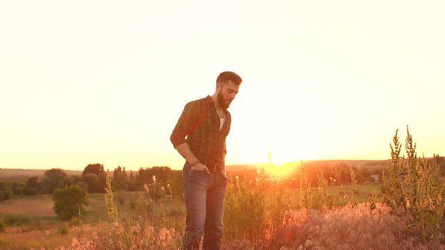 Beautiful bearded man stands on the backdrop of the setting sun in a field in the countryside, slow motion.