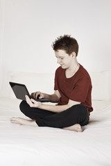 Teen boy sitting on a bed using a laptop computer.