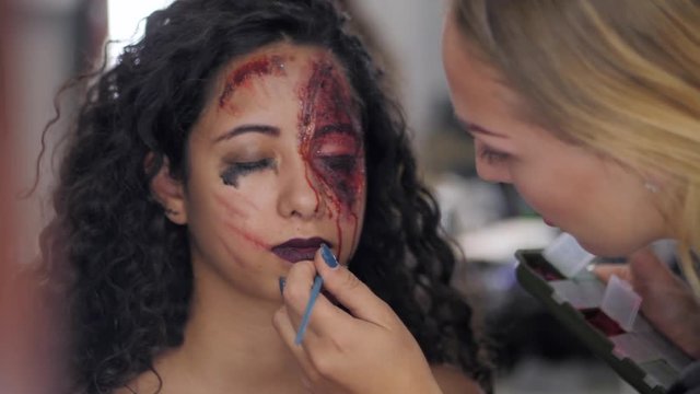 Make-up artist make the girl halloween make upin studio.Halloween face art.Woman applies on professional greasepaint on the face of spanish girl.War-paint with blood, scars and wounds.Slow motion.