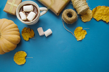 Cup of hot cocoa with marshmallows, gifts, packing paper, dry leaves on a blue table. top view, flat lay, autumn concept, space for text