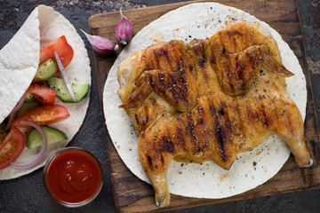 Rustic wooden serving board with roasted chicken tabaka on tortilla bread, view from above