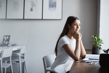 Young lady deciding on her career path sitting in bright coworking space studying. Looking outside smiling.