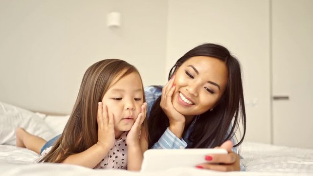Smiling pretty asian woman lying on bed with her young daughter and using smartphone