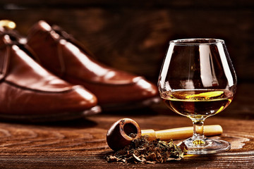 Glass of cognac and pipe with tobacco with glance shoes defocused on second background on wooden table