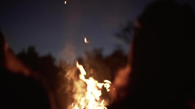 silhouettes of two girls sitting in front bonfire with flame sparks and smoke rising to sky near wood slow motion closeup