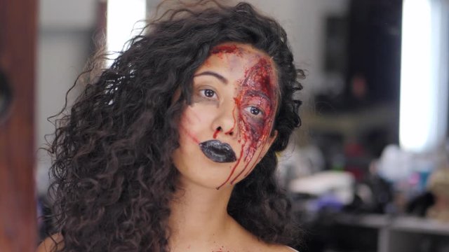 Scary portrait of young girl with Halloween blood makeup. Beautiful latin woman with curly hair looking into mirror reflection in the dressing room. Preparation for celebrating. Slow motion.