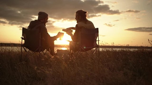Two men sitting in camping chairs in a field watching sunset pouring tea from thermal mugs.