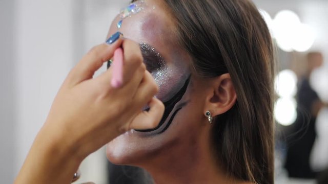 Makeup artist paints greasepaint for Halloween in studio. Woman drawing a glamorous skull with rhinestones and sequins on a beautiful young girl with long hair. Slow motion.