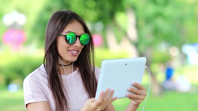 Young woman sits on bench in city park and communicates via tablet pc. Attractive woman in glasses with white tablet computer communicates through skype. Female with tablet pc, she is in a merry mood